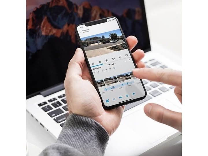 Hands holding a phone over a laptop, with a security camera app open to playback of a driveway and a timeline of saved clips.